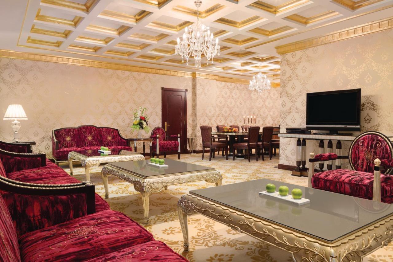 Grand Regency Doha, Trademark Collection By Wyndham Luaran gambar The photo depicts an elegantly designed living room. The room features luxurious red sofas with ornate designs and metallic gold accents, arranged around glass-topped coffee tables that have intricate bases. A large, modern flat-screen television is 