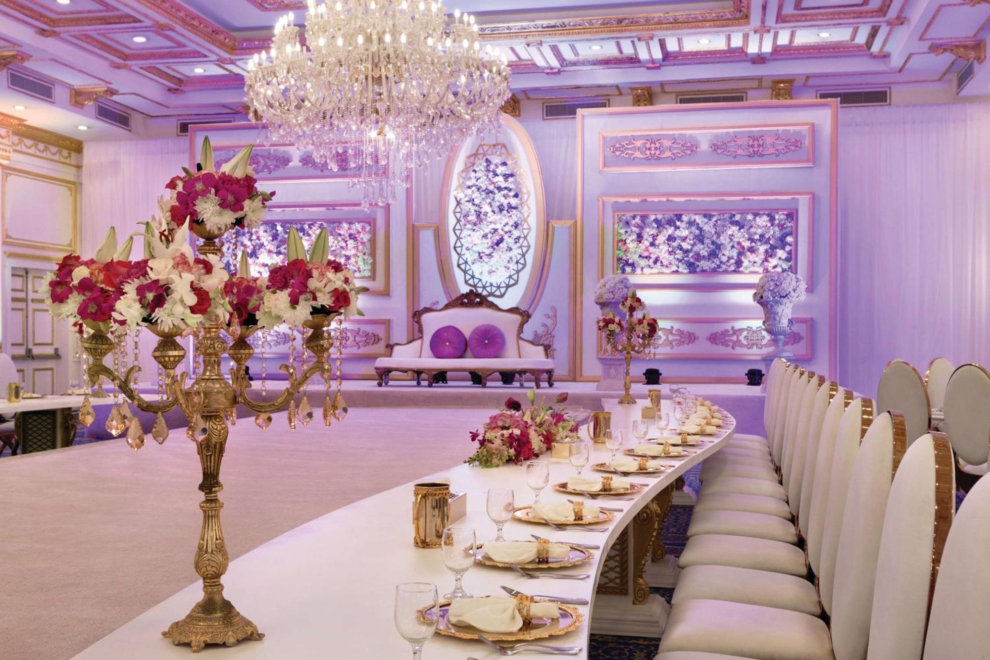 Grand Regency Doha, Trademark Collection By Wyndham Luaran gambar The photo displays an elegantly decorated banquet or dining hall. It features a long, curved table set for a formal event, with fine china and floral centerpieces. Luxurious decorations include a grand chandelier hanging from the ceiling, and the wal