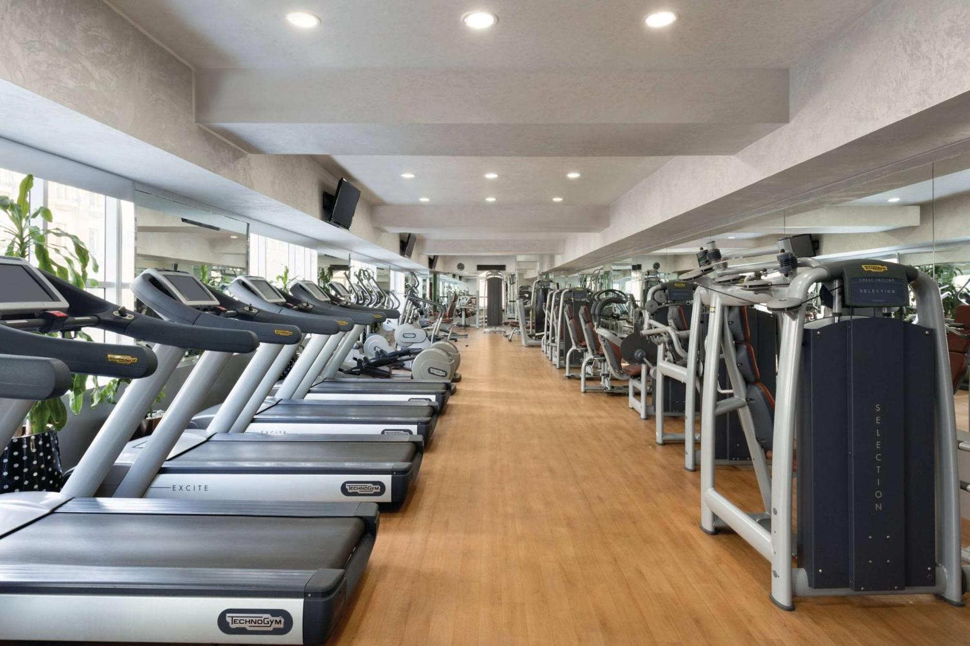 Grand Regency Doha, Trademark Collection By Wyndham Luaran gambar The photo shows the interior of a gym or fitness center. It features a row of modern treadmills lined up against a wall, with a polished wooden floor. The space is brightly lit with recessed ceiling lights, and there are large mirrors on the walls, e