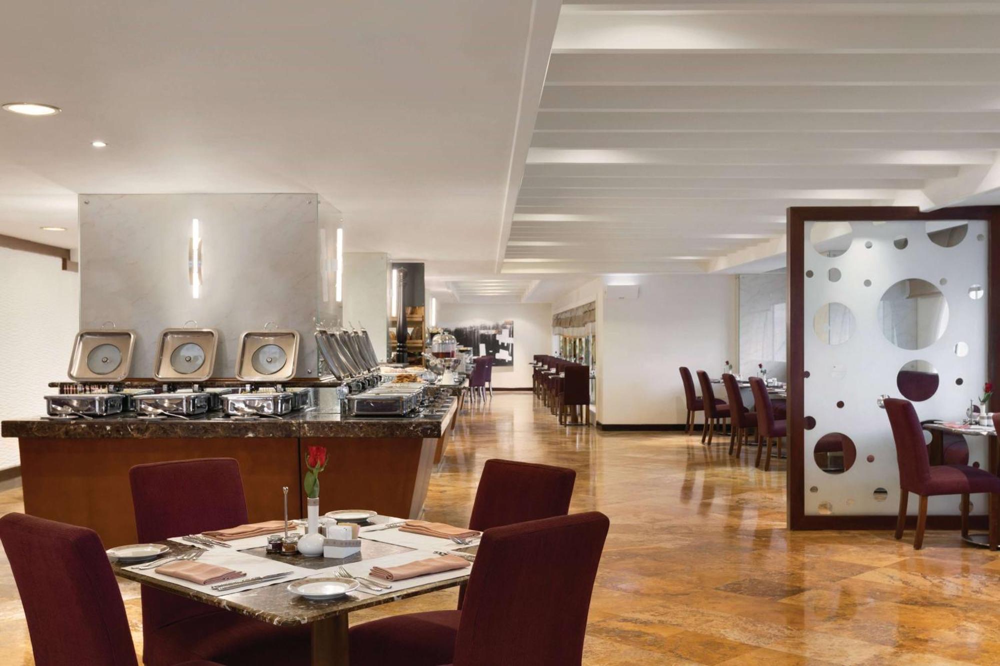 Grand Regency Doha, Trademark Collection By Wyndham Luaran gambar The photo shows a modern restaurant interior. There are several tables with maroon chairs arranged throughout the space. One table is set with a small vase containing a flower and dining utensils. Along one side, there is a buffet area with a sleek c