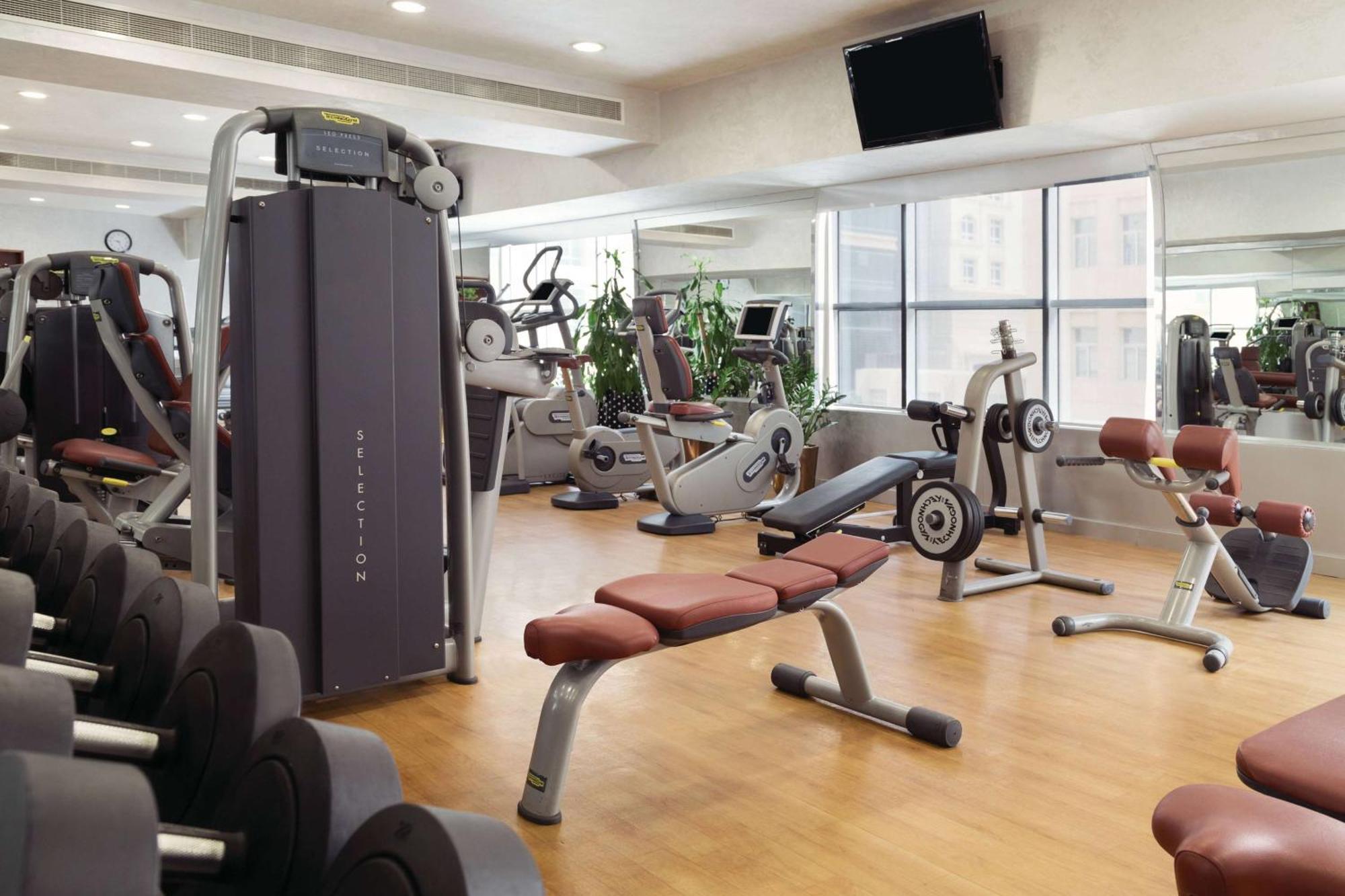 Grand Regency Doha, Trademark Collection By Wyndham Luaran gambar The photo shows a fitness gym with various exercise equipment. There are weight machines, benches, and cardio machines such as stationary bikes and treadmills. The space has large windows allowing natural light to enter, and there are mirrors on the 
