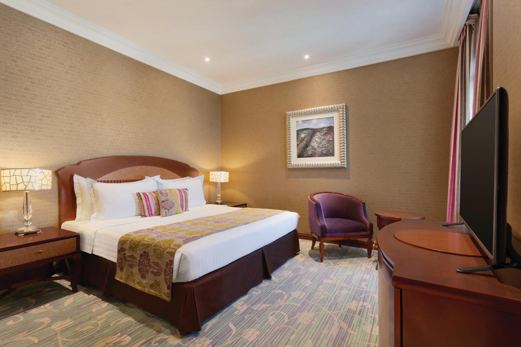 Grand Regency Doha, Trademark Collection By Wyndham Luaran gambar The image shows a hotel room that features a cozy and elegant design. There's a large bed with white sheets and colorful decorative pillows. Next to the bed, there are nightstands with lamps. A comfortable armchair is positioned in the corner, and a 