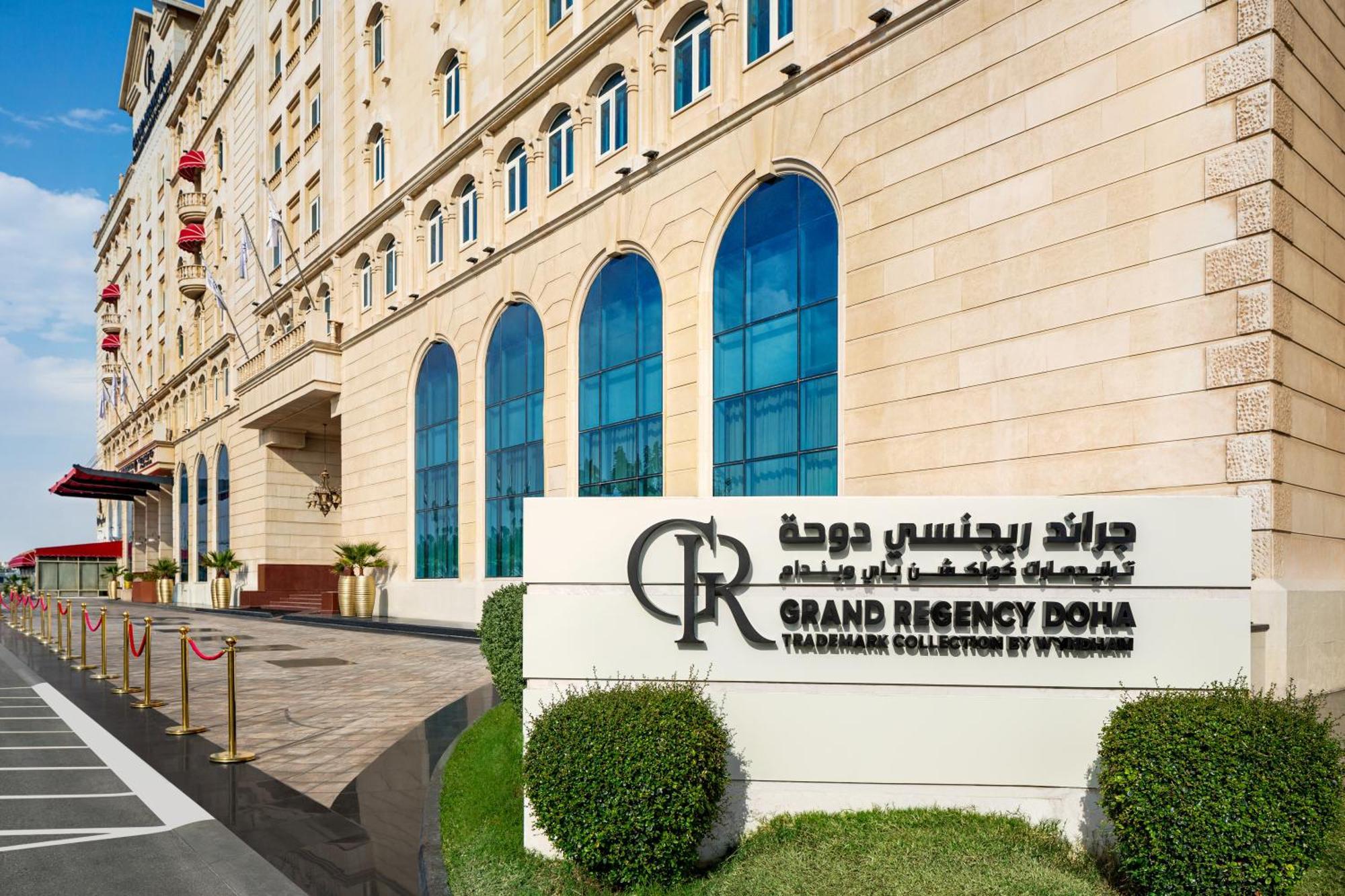 Grand Regency Doha, Trademark Collection By Wyndham Luaran gambar The photo shows the entrance of the Grand Regency Doha, a luxury hotel. The building features a distinctive architectural style with large arched windows. There is a prominent sign featuring the hotel's name in both English and Arabic. The surroundin