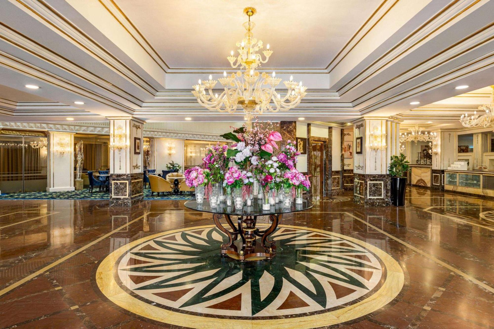 Grand Regency Doha, Trademark Collection By Wyndham Luaran gambar The photo shows a luxurious hotel lobby featuring an elegant chandelier hanging from the ceiling. In the center, there is a large round table adorned with an arrangement of vibrant flowers. The floor showcases intricate marble patterns, and the overa