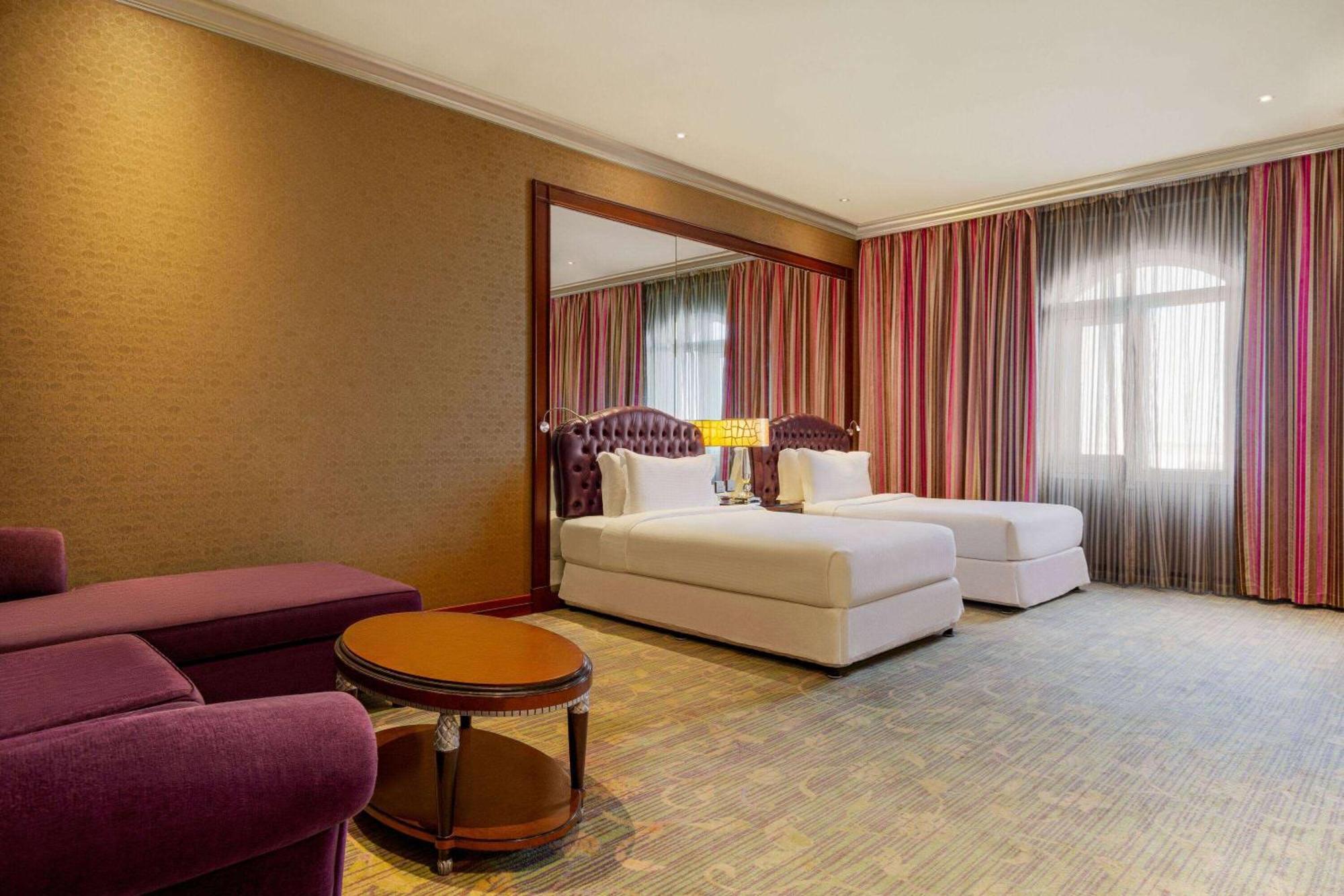 Grand Regency Doha, Trademark Collection By Wyndham Luaran gambar The photo shows a hotel room featuring two beds with white bedding, a large mirror on one wall, and a window dressed with sheer and curtain panels. There is a small round table in the center of the room and a plush purple armchair nearby. The overall