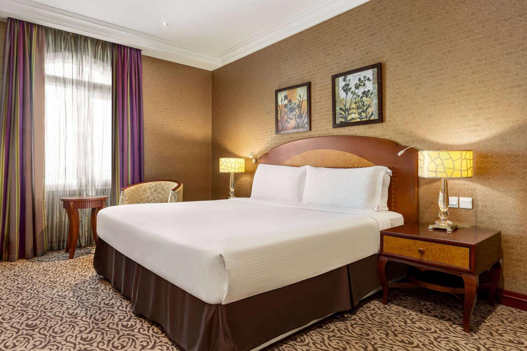 Grand Regency Doha, Trademark Collection By Wyndham Luaran gambar The photo displays a comfortable hotel room featuring a large bed with white linens and a brown bed skirt. The room has a warm color palette, with textured brown walls and patterned carpet. There are two bedside lamps providing soft lighting next to 