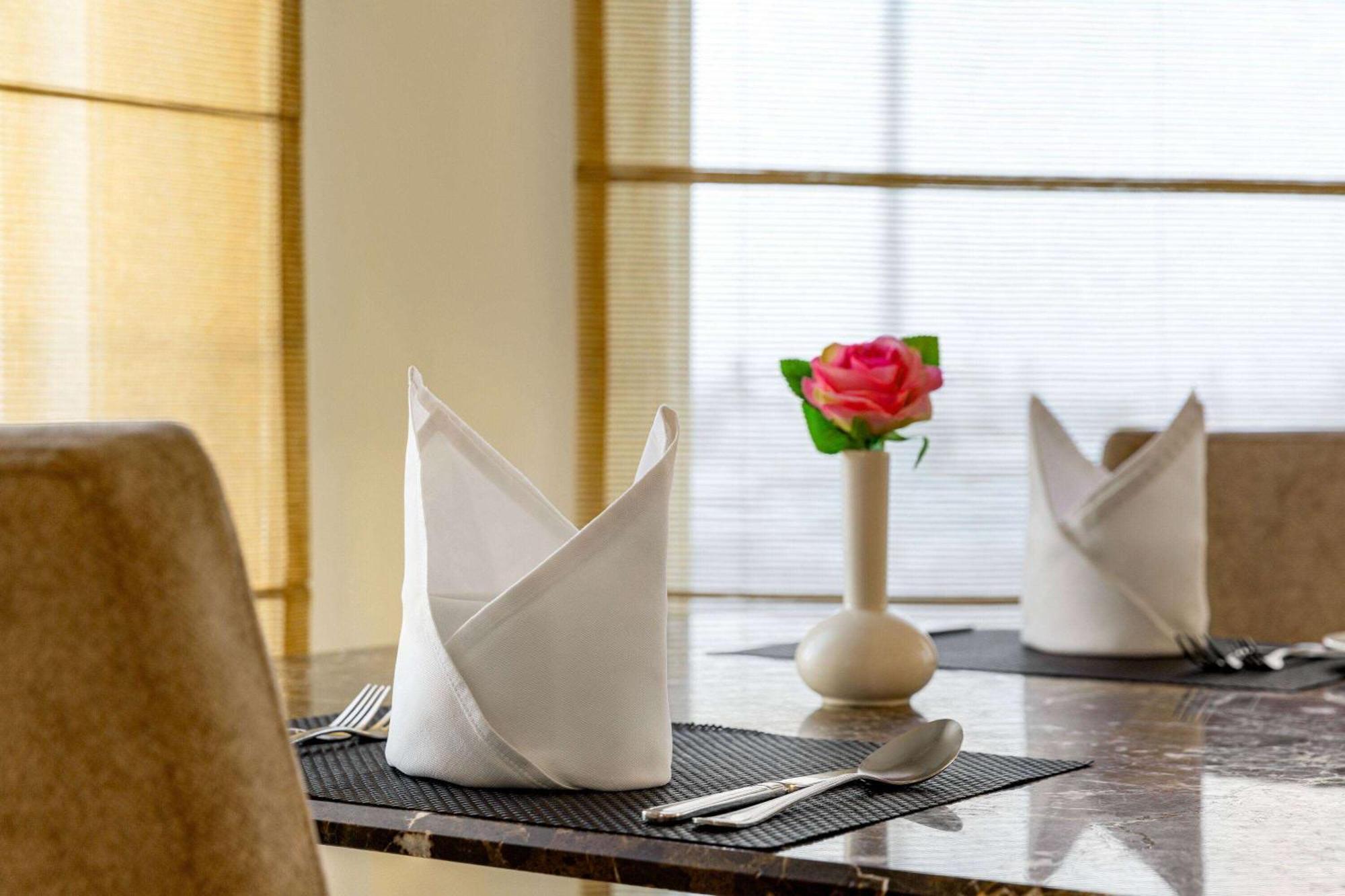 Grand Regency Doha, Trademark Collection By Wyndham Luaran gambar The photo shows a neatly arranged dining table. On the table, there are two cloth napkins folded in a decorative way, one at each place setting. Silverware, including a knife and fork, is placed next to each napkin. In the center of the table, there 