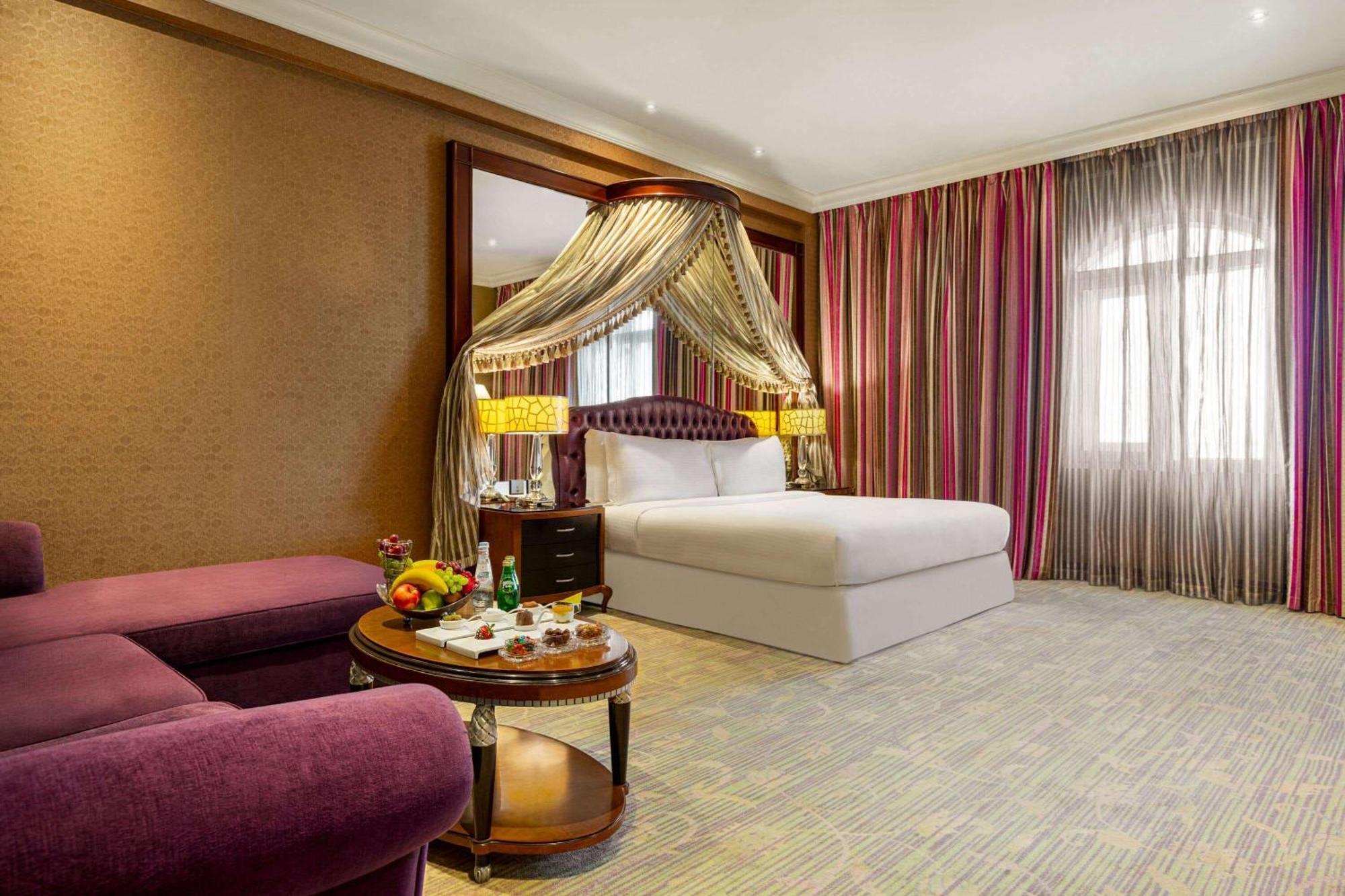 Grand Regency Doha, Trademark Collection By Wyndham Luaran gambar The photo shows a luxurious hotel room featuring a king-sized bed with a decorative canopy. The room is elegantly designed with a mix of rich colors and textures. To the left, there's a plush purple seating area with a small round table that displays